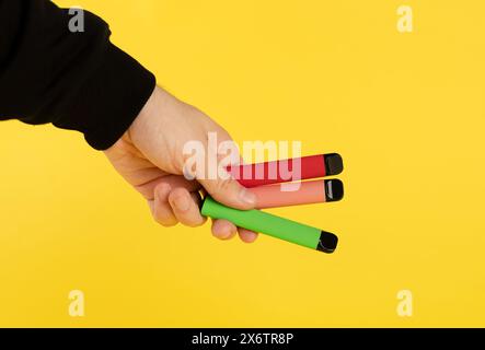 Elektronische Einweg-Zigaretten verschiedener Geschmacksrichtungen in der Hand auf einem gelben Hintergrund. Das Konzept von modernem Rauchen, Verdampfen und Nikotin. Stockfoto