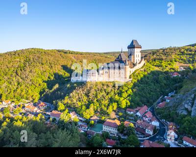 Ein atemberaubender Blick aus der Vogelperspektive auf das Schloss Karlstejn auf einem Hügel, umgeben von üppigen grünen Wäldern und einem malerischen Dorf, unter klarem blauen Himmel am Morgen. Tschechien Stockfoto