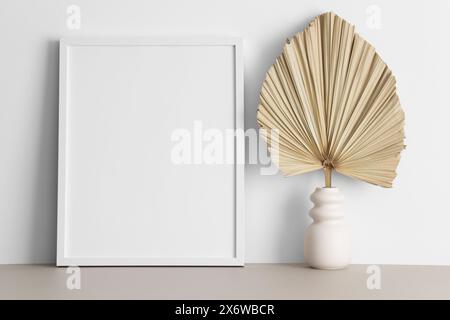 Weißes Rahmenmockup mit Palmblattdekoration auf dem beigefarbenen Tisch. Stockfoto