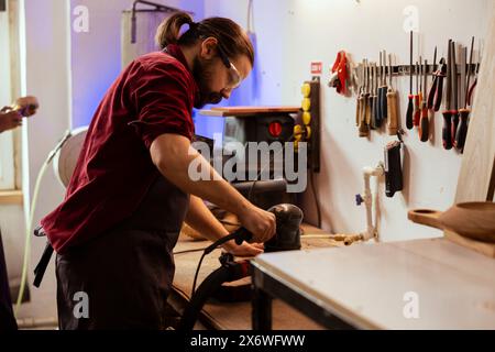 Zimmermann mit Winkelschleifer auf Holzblock zum Glätten von Oberflächen durch Abrieb mit Schleifpapier. Holzbearbeiter mit Schwingschleifer-Ausrüstung für die Möbelmontage im Atelier Stockfoto