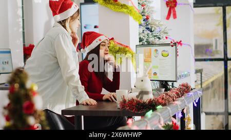 Angestellter mit Weihnachtsmütze, der im weihnachtlich verzierten Büro arbeitet und um Hilfe von einem Kollegen bittet. Mitarbeiter, die mit Kollegen im Weihnachtsgeschäft die Umsatzzahlen des Unternehmens betrachten, schmücken den Arbeitsplatz, die Handkamera Stockfoto