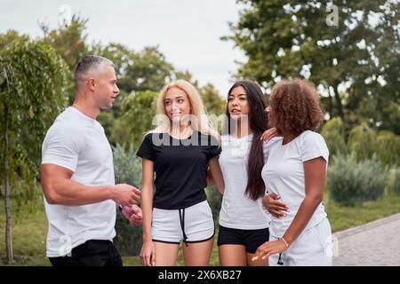 Hübscher junger männlicher Trainer, der mit verschiedenen Frauen in Sportbekleidung im Park spricht. Multiethnische, fitte Freundinnen, die während des Trainings in der Natur dem Coach zuhören. Stockfoto