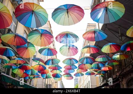 Farbenfrohe Regenschirme schmücken die Straße und sorgen für ein lebendiges Display vor blauem Himmel. Viele Regenbogenschirme tragen zur festlichen Straßendekoration bei, besonders während des Pride Month. Stockfoto