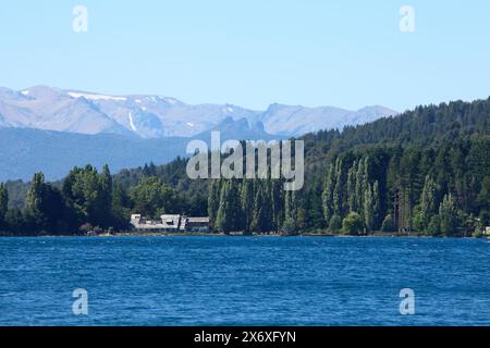 Dicht grüner Wald auf einem Berghang in der Nähe eines blauen Sees mit einem alten Gebäude an der Küste und Berggipfeln in der Ferne, Bariloche Nationalpark Stockfoto
