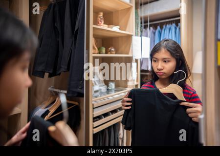 Hübsche indonesische Teenager-Mädchen, die Kleidung aussuchen und denken, während sie in den Spiegel im Schrank zu Hause schauen. Asiatische Frau entscheidet, welches Hemd sie tragen soll Stockfoto