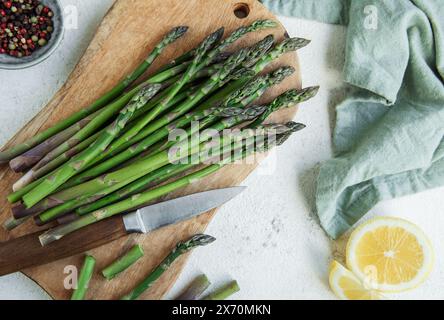 Frische, grüne Spargelspeere werden auf einem hölzernen Schneidebrett neben einem scharfen Küchenmesser mit einer halben Zitrone und einer Schüssel Pfeffer ne ordentlich angeordnet Stockfoto