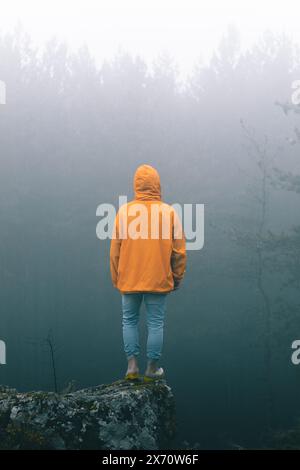 Bulgarien - 05 22 2024: Mann mit leuchtender orangefarbener Jacke und Jeans, der am Felsenrand steht und auf das helle Licht über den Bäumen blickt Stockfoto