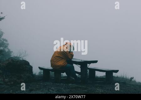 Bulgarien - 22 05 2024: Ein trauriger Mann mit orangefarbener wasserdichter Jacke und Jeans sitzt auf einer Bank auf einem Hügel, während er regnet und seinen Kopf hält. Stockfoto