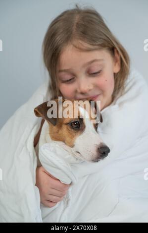 Das kleine kaukasische Mädchen umarmt einen Jack Russell Terrierhund in eine Decke gewickelt. Vertikales Foto. Stockfoto