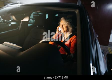 Eine ältere Frau, die in einem Auto sitzt, das in einer Tiefgarage geparkt ist und lächelt, während sie am Telefon spricht. Stockfoto