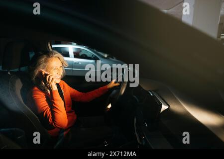 Eine ältere Frau, die in einem Auto sitzt, das in einer Tiefgarage geparkt ist und lächelt, während sie am Telefon spricht. Stockfoto