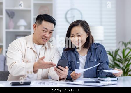 Lächelndes Paar, das Finanzdokumente überprüft und zu Hause ein Smartphone verwendet, um Teamarbeit und Finanzplanung zu demonstrieren Stockfoto