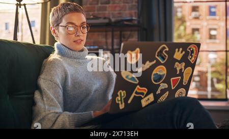 Eleganter Laptop für Frauen mit diversen LGBT- und Lifestyle-Aufklebern auf der Rückseite. Junge kreative Frau sitzt auf einer Couch, tippt, surft im Internet und schaut zu Hause in sozialen Medien. Stockfoto