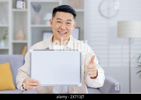 Fröhlicher Mann, der ein leeres Whiteboard präsentiert und die Daumen nach oben zeigt, während er in einem hellen, modernen Wohnzimmer steht Stockfoto