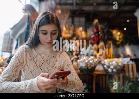 Ein fesselndes, farbenfrohes Mädchen, das die Weihnachtsdekoration bewundert und ihr Smartphone durchsucht. Stockfoto