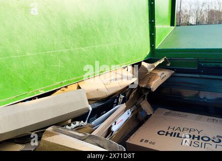 Paris, Frankreich - 1. März 2024: Ein grüner Müllcontainer gefüllt mit heruntergekommenen Kartonschachteln, darunter eine mit Happy Childhood beschriftete im Hintergrund zeigt eine Umgebung im Freien mit Bäumen und einem klaren Himmel. Stockfoto