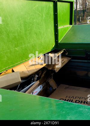 Paris, Frankreich - 1. März 2024: Ein grüner Müllcontainer gefüllt mit heruntergekommenen Kartonschachteln, darunter eine mit Happy Childhood beschriftete im Hintergrund zeigt eine Umgebung im Freien mit Bäumen und einem klaren Himmel Stockfoto