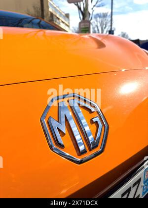 Freiburg - 2. März 2024: Nahaufnahme des MG-Logos auf der Motorhaube eines hellen orangen Elektroautos. Die lebendige Farbe und das glänzende Emblem zeichnen sich durch einen verschwommenen urbanen Hintergrund und einen klaren blauen Himmel aus Stockfoto