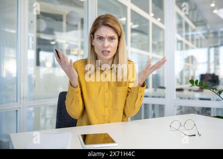 Blonde Geschäftsfrau mit gelbem Hemd, die frustriert und verwirrt am Schreibtisch sitzt. Konzept von Stress und Herausforderungen am Arbeitsplatz. Stockfoto