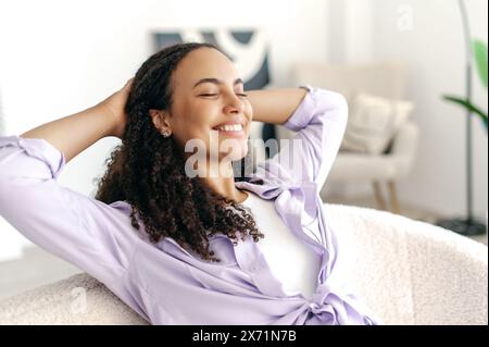 Glückliche entspannte, ruhige, lockige, stilvolle Frau, die eine Pause von der Online-Arbeit macht, sich mit den Händen hinter dem Kopf ausruht und die Augen schließt, während sie auf einem Sofa sitzt und fröhlich lächelt Stockfoto