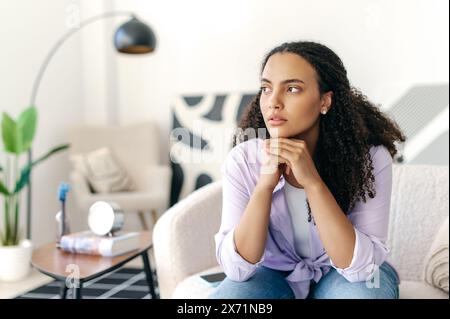 Besorgte depressive junge brasilianische oder hispanische Frau mit lockigen Haaren, die allein auf einem Sofa sitzt, an Einsamkeit leidet, sich gestresst fühlt, müde ist, über schlechte Nachrichten nachdenkt, Probleme in Beziehungen Stockfoto