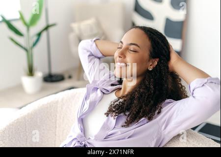 Entspannte, ruhige, lockige, stilvolle Frau, die eine Pause von der Online-Arbeit macht, sich mit den Händen hinter dem Kopf ausruht und die Augen schließt, während sie lächelnd auf einem Sofa sitzt Stockfoto