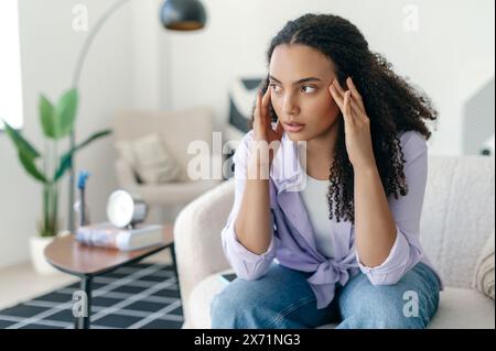 Depressiv frustrierte junge brasilianische oder hispanische Frau, allein auf einem Sofa sitzend, unter Kopfschmerzen, Migräne, Stress, Müdigkeit, denkt über schlechte Nachrichten nach, Probleme in Beziehungen Stockfoto
