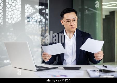 Ein reifer asiatischer Mann an seinem Schreibtisch in einem modernen Büro, der Dokumente aufmerksam überprüft. Professionelles Ambiente mit Laptop und Papieren. Stockfoto