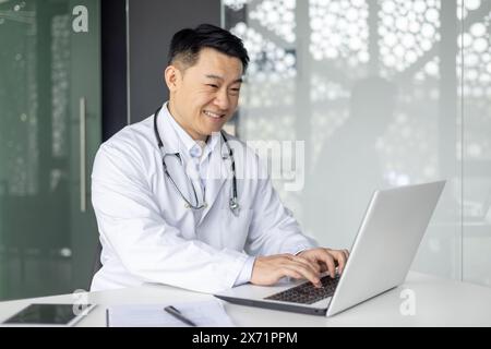 Asiatischer reifer Arzt im weißen Labormantel, der im Büro an einem Laptop arbeitet, lächelt und fröhlich und professionell aussieht. Stockfoto