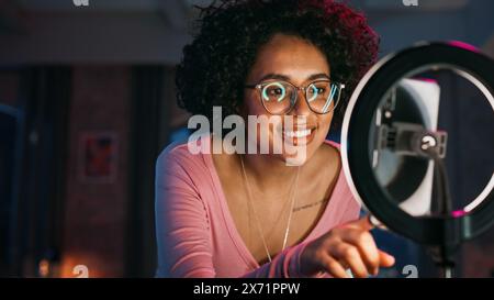 Schöne junge Frau richtet Studio Lighting und Smartphone ein, um eine Tanzroutine auf Video für soziale Medien aufzunehmen. Stilvolles schwarzes Mädchen in Brille streamt ihre Choreografie im Internet von zu Hause aus. Stockfoto