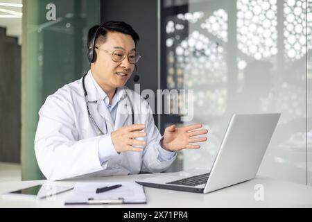 Asiatischer reifer Arzt mit Laptop für eine Online-Konsultation, mit Headset in einer modernen Arztpraxis. Stockfoto