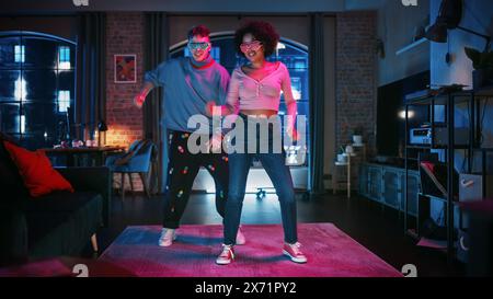 Schönes stilvolles multiethnisches Paar in legeren Outfits und futuristischen Neon Gllowing Brille, Tanz und genieße das Leben zu Hause in Loft Apartment. Aufnahme lustiger viraler und aktiver Videos für soziale Medien. Stockfoto