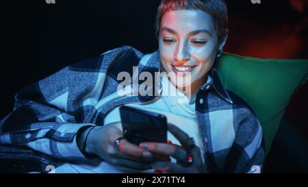 Nahaufnahme Porträt einer Happy Young Female mit kurzen Haaren, die auf einer Couch zu Hause liegen, während sie ein Smartphone verwenden. Schöne, vielfältige Frau, die im Internet surft, soziale Medien durchsucht und mit Freunden chatten kann. Stockfoto