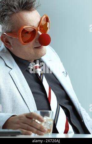 In einem leichten Anzug und einer gestreiften Krawatte trägt der Mann übergroße orangene Gläser und eine rote Clown-Nase, während er ein Whiskey-Glas hält. Sein dummes Verhalten hig Stockfoto