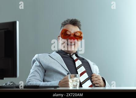 Ein Mann, in einem leichten Anzug und einer gestreiften Krawatte, trägt eine komische orange Brille und eine rote Clownnnase, hält ein Glas Whiskey. Sein lächerlicher Blick unterstreicht t Stockfoto