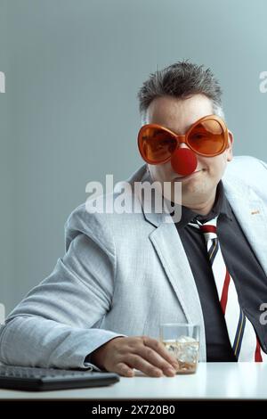 Ein Mann mittleren Alters in einem leichten Anzug und einer gestreiften Krawatte trägt eine übergroße orange Brille und eine rote Clownnnase, die ein Whiskyglas hält. Sein verspieltes, lächerliches Stockfoto