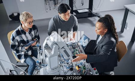 Schwarzer Lehrer mit Laptop und zwei verschiedene Schüler analysieren und diskutieren den futuristischen Roboterarm während der Lektion. Dozent hilft beim Projekt. Informatikkonzept. Hochwinkelansicht. Stockfoto
