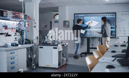 Ein vielfältiges Team aus zwei Schülern steht in der Nähe eines großen Bildschirms mit Tablet und Laptop, während sie über Programmierung und Technologien diskutieren. Ausbildung und Studium im modernen Technischen Hochschulkonzept. Stockfoto