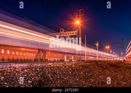 Der Zug fährt bei Nacht vorbei und hinterlässt leichte Wege in verschiedenen Farben. Szene mit beleuchteten Signalen, Hochspannungsmasten und Kontaktdrähten. Kies von t Stockfoto