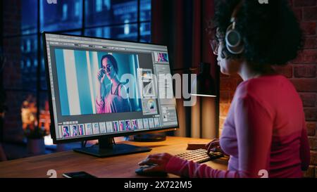 Junge kreative Frau in Kopfhörern mit Computer in stilvollem Loft Apartment am Abend. Grafikdesigner arbeitet von zu Hause aus, bearbeitet Modefotos für den Online-Shop. Blick auf die Stadt vom großen Fenster. Stockfoto