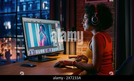 Junge kreative Frau in Kopfhörern mit Computer in stilvollem Loft Apartment am Abend. Grafikdesigner arbeitet von zu Hause aus, bearbeitet Modefotos für den Online-Shop. Blick auf die Stadt vom großen Fenster. Stockfoto