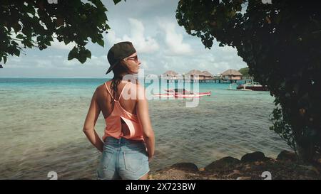 Eine Frau in lässiger Kleidung steht an einem Sandstrand am Meer und blickt an einem sonnigen Tag unter dem klaren Himmel auf das Wasser. Stockfoto