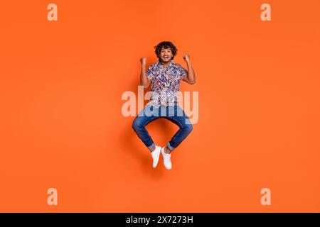 Foto in voller Länge von gutaussehendem jungen Mann springen heben Fäuste gekleidet stilvolle bunte Kleidung isoliert auf orangefarbenem Hintergrund Stockfoto