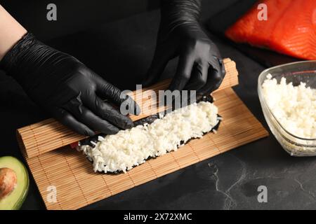 Koch in Handschuhen, die Sushi-Rolle an einem dunklen, strukturierten Tisch einwickeln, Nahaufnahme Stockfoto