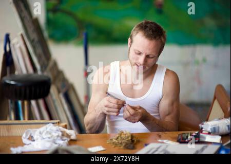 Junger Erwachsener Mann genießt einen Joint junger Erwachsener Mann genießt einen Joint mit Haschisch / Pot, während er auf einem Tisch in einem Kunststudio sitzt. Rotterdam, Niederlande. Rotterdam Kraakpand Merwehaven Zuid-Holland Nederland Copyright: XGuidoxKoppesxPhotox Stockfoto