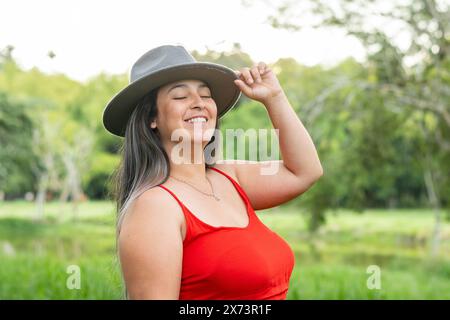 latina lächelt glücklich, als sie die Augen schließt und ihren grauen Hut in der Hand hält Stockfoto