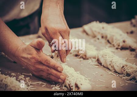 Ein Bäcker legt Käse auf rohe Teigbrötchen auf das Blech in der Bäckerei. Vintage-Stil mit Körnung. Stockfoto