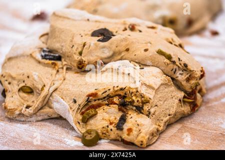 Roher Brotteig mit Oliven, Tomaten und Kräutern, auf dem Holzarbeitstisch, für die weitere Verarbeitung in einer Bäckerei vorbereitet. Stockfoto