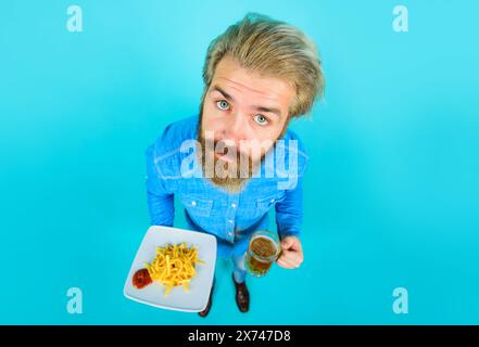 Fastfood. Bärtiger Mann mit Teller leckerer Pommes frites und Ketchup halten einen Becher Bier. Köstliches Essen. Gutaussehender Kerl trinkt Bier und isst Kartoffelfritten. Stockfoto