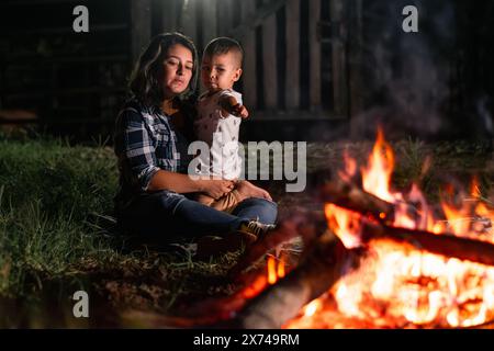 Eine junge lateinfrau, die ihr Kind trägt, nachts vor einem Lagerfeuer sitzt, auf das neugierige Kind zeigt Stockfoto